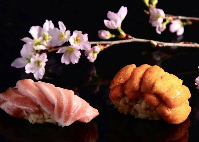 Two pieces of refined sushi at Jukusei Sushi Wagokoro, set against a black backdrop and cherry blossoms.